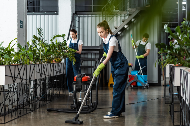 Professionelles Reinigungsteam in Uniform bei der sorgfältigen Reinigung eines modernen Büros. Eine Mitarbeiterin im Vordergrund staubsaugt den Boden, während im Hintergrund weitere Kräfte Wisch- und Pflegearbeiten durchführen.