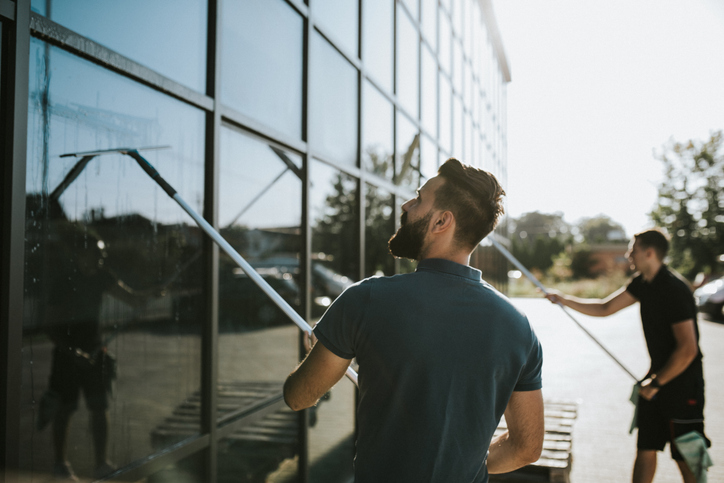 Zwei professionelle Fensterreiniger in Arbeitskleidung säubern bei Sonnenschein die große Glasfassade eines modernen Gebäudes mit langstieligen Abziehern.
