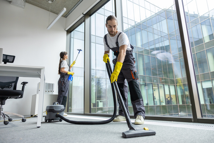 Zwei professionelle Reinigungskräfte in Arbeitsuniform reinigen ein modernes Büro mit großer Fensterfront. Ein Mitarbeiter im Vordergrund saugt gründlich den Teppichboden, während seine Kollegin im Hintergrund die Fenster reinigt.