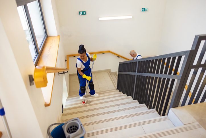 Professionelle Reinigungskraft in Uniform wischt sorgfältig die Steintreppe in einem hellen, modernen Treppenhaus.