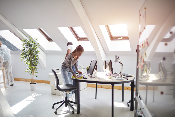 Eine Mitarbeiterin reinigt sorgfältig die Oberfläche eines Schreibtisches und die Tastatur in einem hellen, modernen Dachgeschoss-Büro mit Oberlichtern.