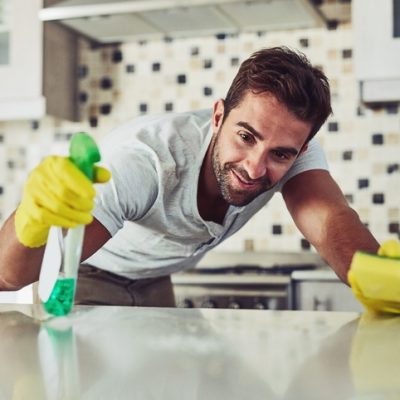 Ein Mann putzt lächelnd mit gelben Handschuhen und einem Schwamm die helle Arbeitsfläche in einer modernen Küche. Tipps zur Vorbereitung auf Unterhalts- und Grundreinigung von der Schlueter Group.